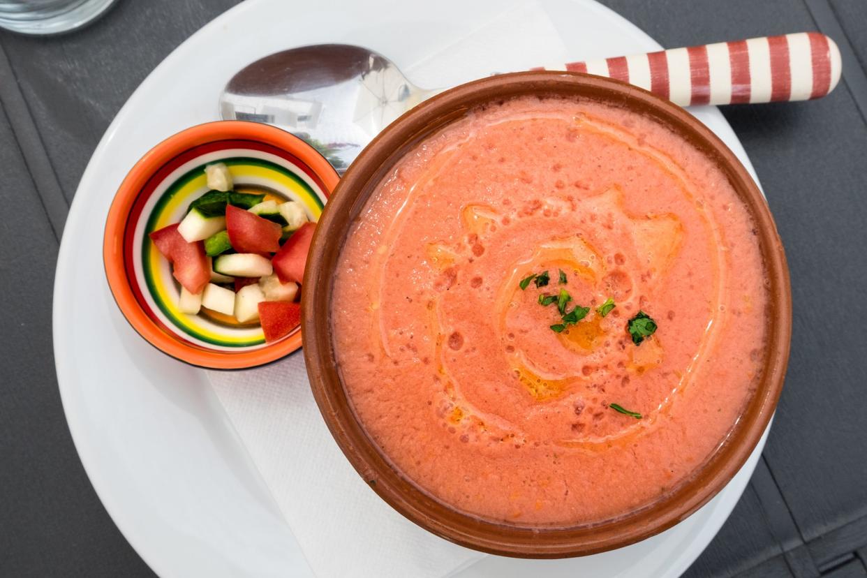 gazpacho with olive oil drizzle, next to spoon and small bowl of chopped vegetables