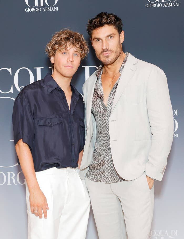 Two men pose together at an event, one in a navy shirt and white pants, the other in a light suit with an unbuttoned shirt