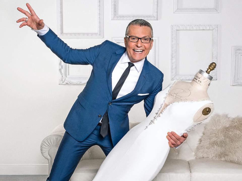 Randy Fenoli holds a mannequin dressed in a white wedding dress in front of a white wall.