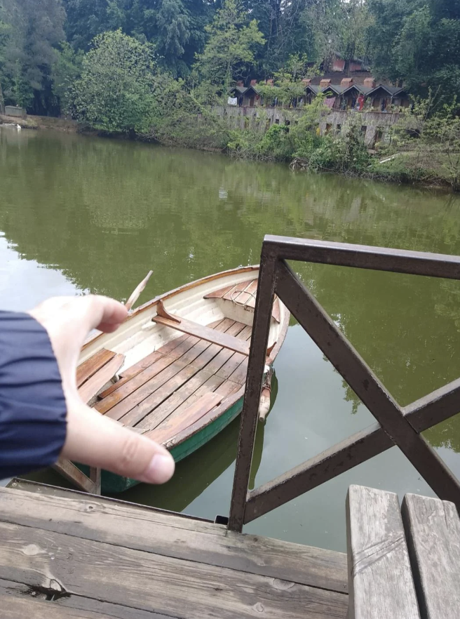 A hand reaches out to reach a rustic wooden rowboat docked next to a wooden platform on a calm, greenish river with forest and cabins visible in the background