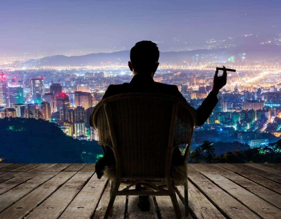 Silhouette of businessman sit on chair and hold a cigar and looking at the city in night.