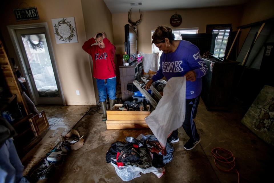 A man puts his hands on his head in a muddy living room as a woman places items into a plastic garbage bag.