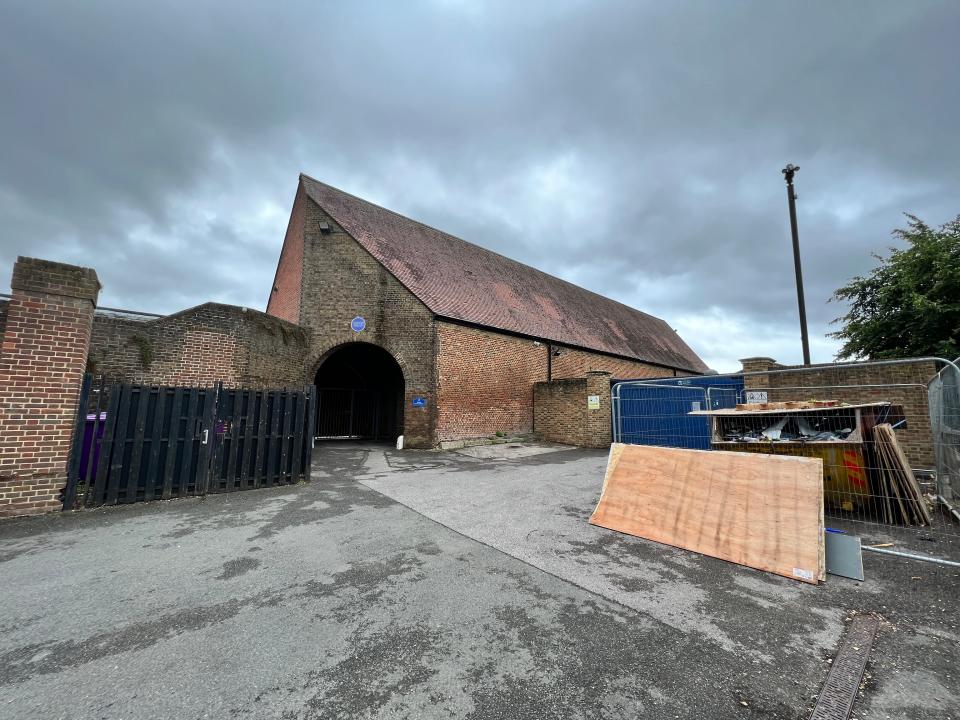 A large windowless brick building with a pointy roof, a converted barn, with a skip and in the front.