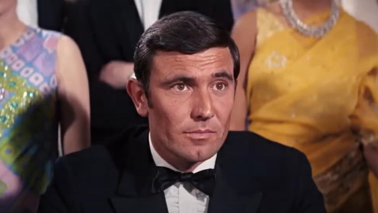  George Lazenby sitting in front of a crowed, dressed in a tuxedo in On Her Majesty's Secret Service. 