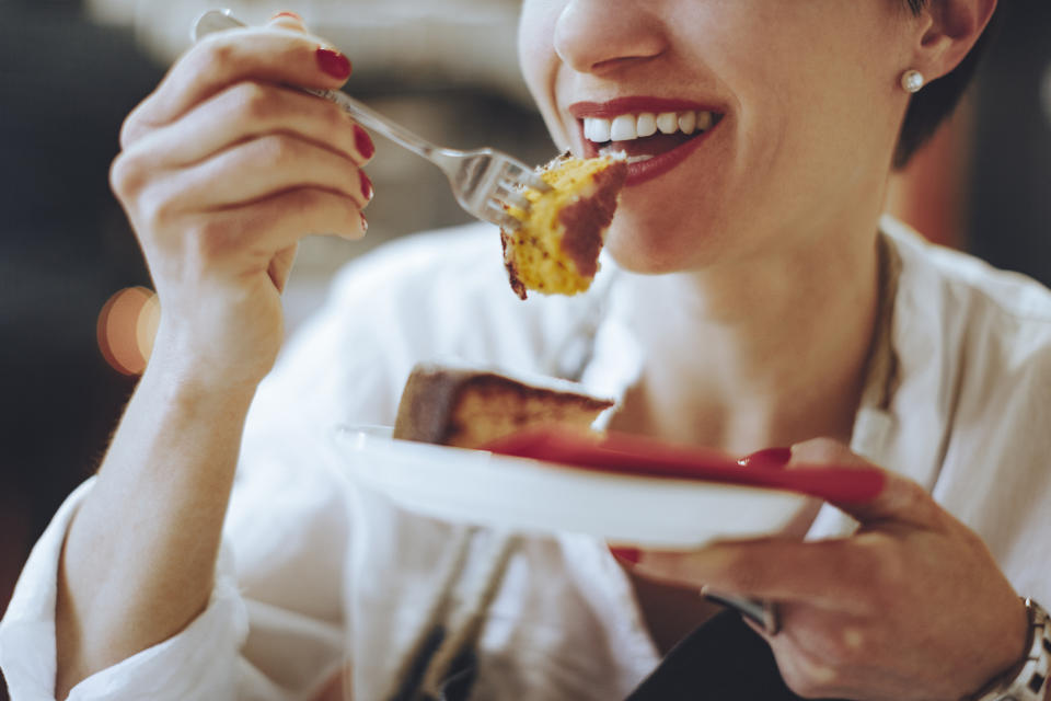 A woman smiles as she enjoys a bite of cake from a plate. She wears a white shirt and has pearl earrings