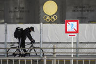 A man wearing a face mask to protect against the spread of the coronavirus rides a bicycle near the Japan National Stadium, where opening ceremony and many other events are planned for postponed Tokyo 2020 Olympics, as engravings in honor of 1964 Tokyo Olympics are seen on the side of the stadium wall behind the fence in Tokyo Monday, Jan. 18, 2021. The Japanese capital confirmed more than 1,200 new coronavirus cases on Monday. (AP Photo/Eugene Hoshiko)