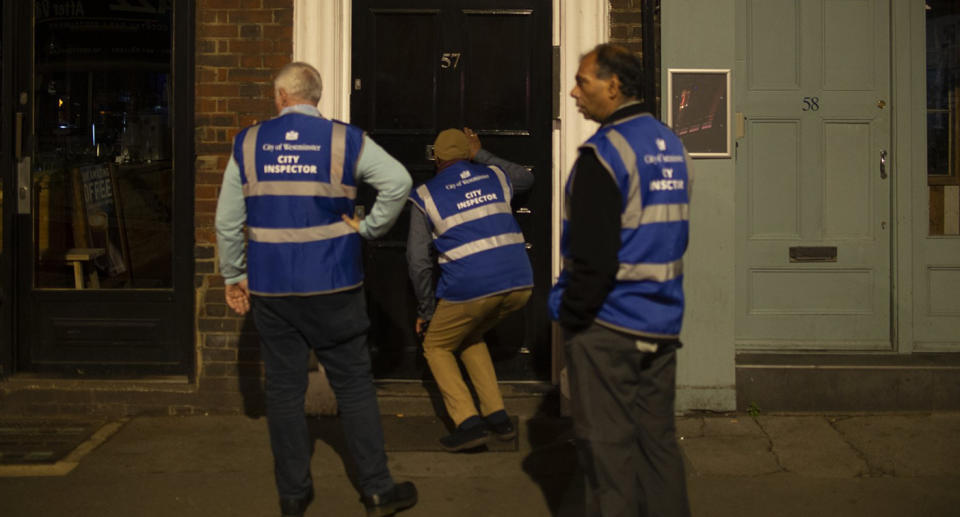 A London city inspector peers through a letterbox slot on the front door of a drinking establishment as two colleagues look on.