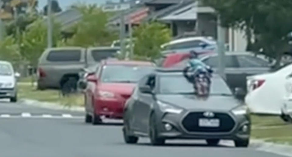 The child is clinging  to the windscreen of the moving grey hatchback, turning his head to the left to watch where the moving car is headed. 