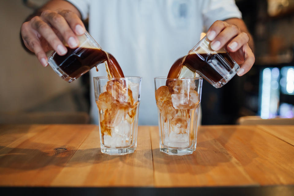 Hands pouring iced coffee into two glasses filled with ice on a wooden table