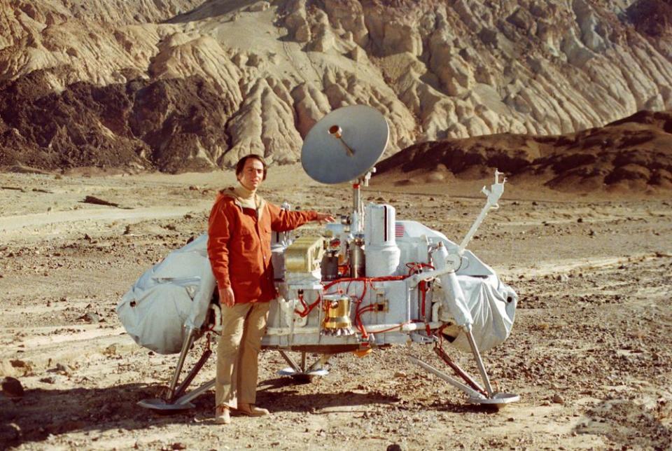 a man in a jacket stands next to a spacecraft in a desert