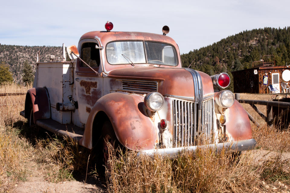 Old rusty fire truck parked in a field with clear skies and trees in the background