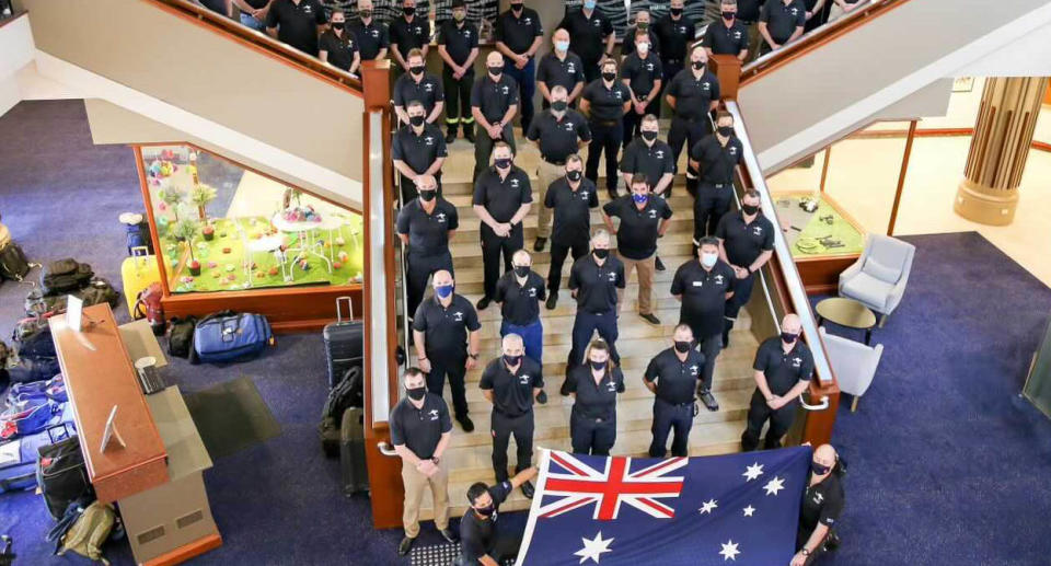 A photo of firefighters at their pre-deployment briefing before flying to Canada. The group poses on a stairway as two firefighters hold an Australian flag at the front. Source: NSW Rural Fire Service