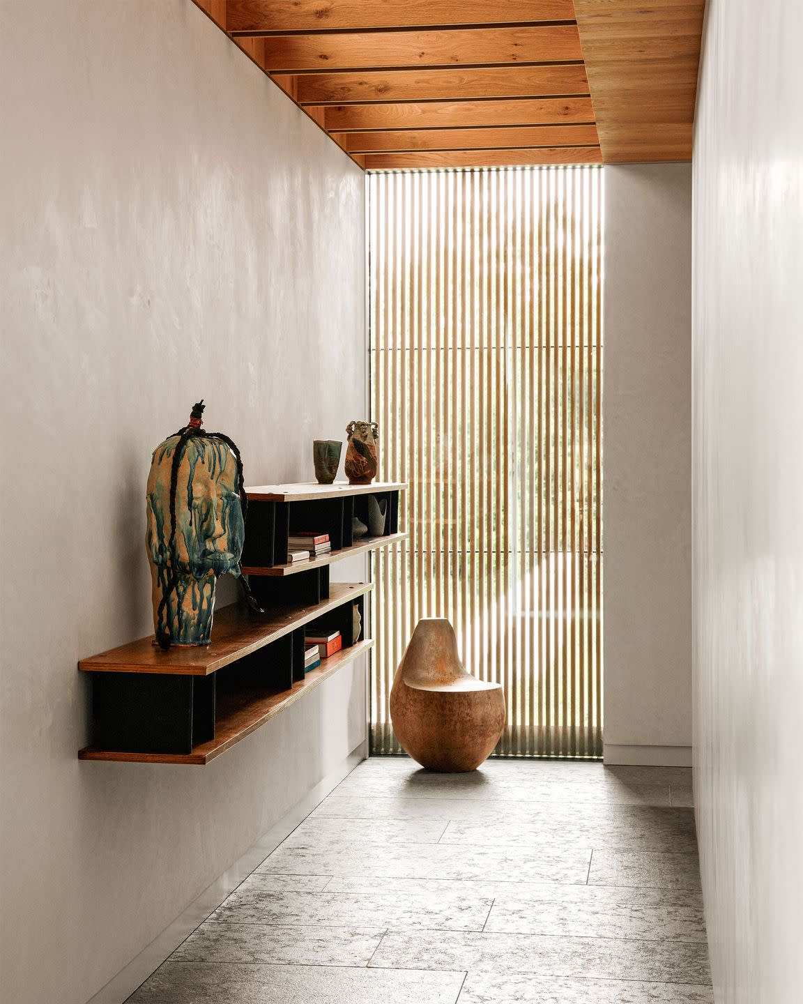 hallway with two floating shelves with compartments of books and objects, sculptures, armless chair, floor to ceiling window with vertical lines, wood beamed ceiling, light textured walls, gray floor tiles