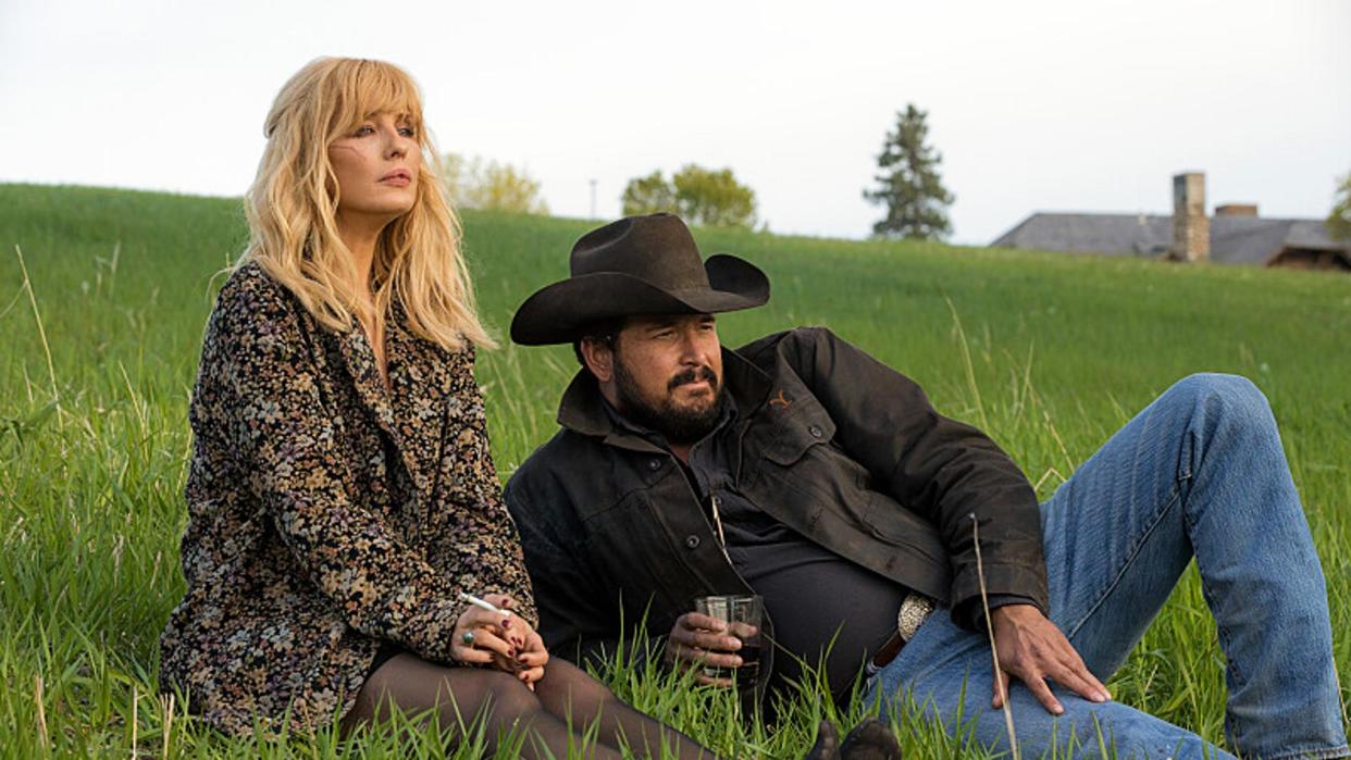  Kelly Reilly as Beth Dutton and Cole Hauser as Rip Wheeler sit in the grass on Yellowstone season 5. 