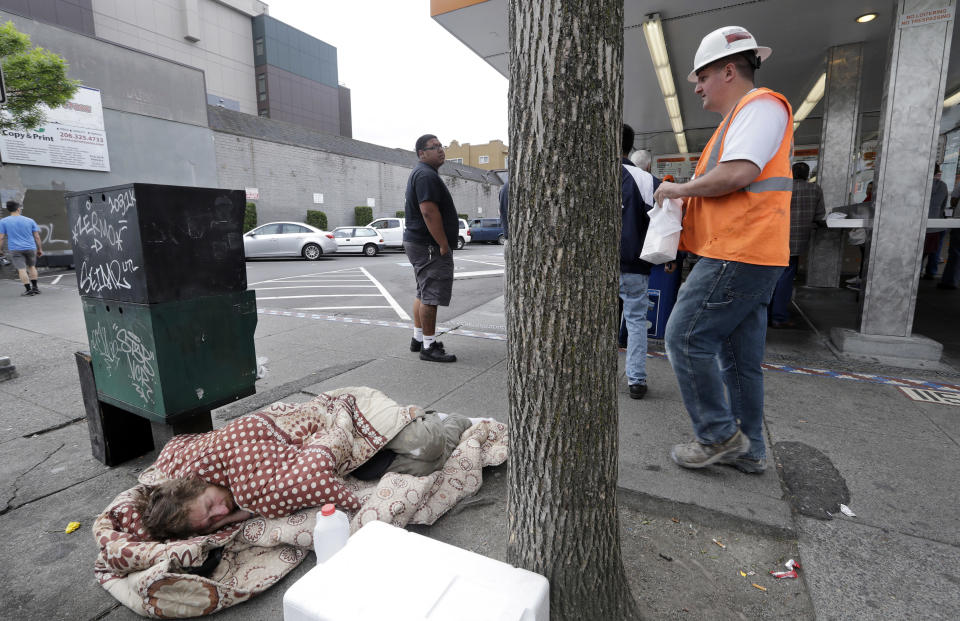 File - In this May 24, 2018, file photo, a man sleeps on the sidewalk as people behind line-up to buy lunch at a Dick's Drive-In restaurant in Seattle. A new federal report says the number of people living on the streets in Los Angeles and San Diego, fell this year, suggesting possible success in those cities' efforts to combat the problem. Meanwhile, homelessness overall was up slightly across the country, including Seattle, although the report did not provide a complete picture of the problem. (AP Photo/Elaine Thompson, File)