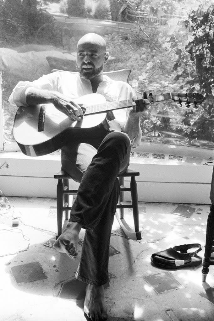 Man sitting, playing guitar barefoot, shoes beside him, relaxed in a sunlit room