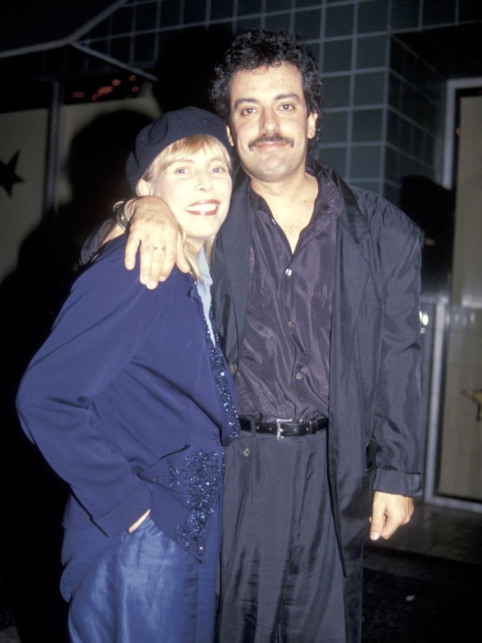 joni mitchell and larry klein smile for a photo while standing and hugging, she wears a beret and blue jacket, he wears a dark suit with a purple collared shirt