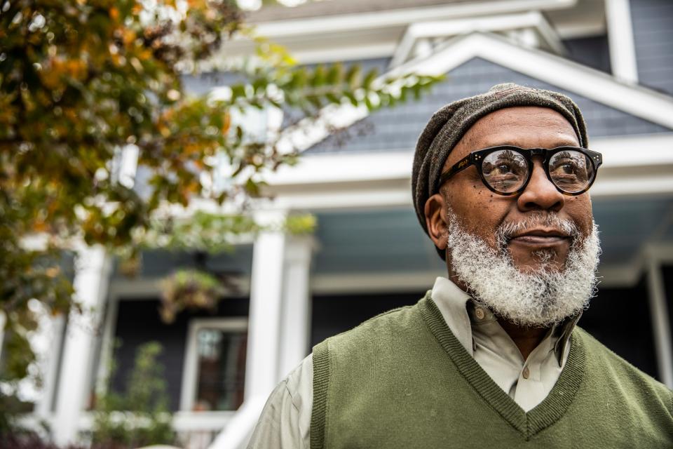 A senior Black man stands in front of a suburban home.