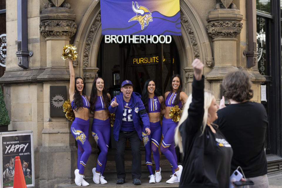 Cheerleaders for the The Minnesota Vikings NFL team pose for pictures with a supporter of the team at a fan interaction event at The Brotherhood Of Pursuits And Pastimes sports bar in Manchester, England, Wednesday, Sept. 28, 2022. A half-dozen NFL teams are aggressively targeting fans in Britain now that they have new marketing rights in the country. They’re signing commercial deals and hiring local media personalities in bids to expand their fanbases and tap international revenue.(AP Photo/Jon Super)