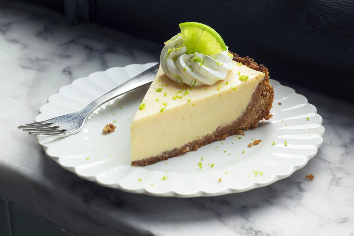 A slice of creamy key lime pie topped with a garnish of whipped cream and lime zest, with a small citrus slice. The crust is made from graham crackers or cookies and the piece of pie is on a white plate with a fork on a marble counter top.