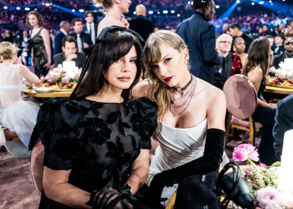 lana del rey and taylor swift pose for a photo while sitting at a table in a crowded room, lana wears all black and taylor wears a white dress with black gloves