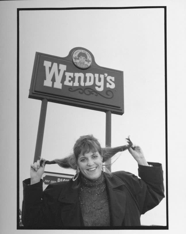 Woman smiling, holding her pigtails under Wendy's sign, mimicking the logo's character. She wears a jacket with a turtleneck