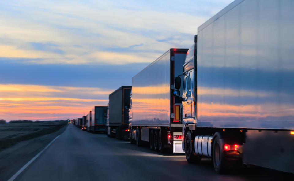 A line of semi-trucks drives on a highway at sunset, reflecting the colorful sky on their sides. The image evokes the theme of travel and transportation