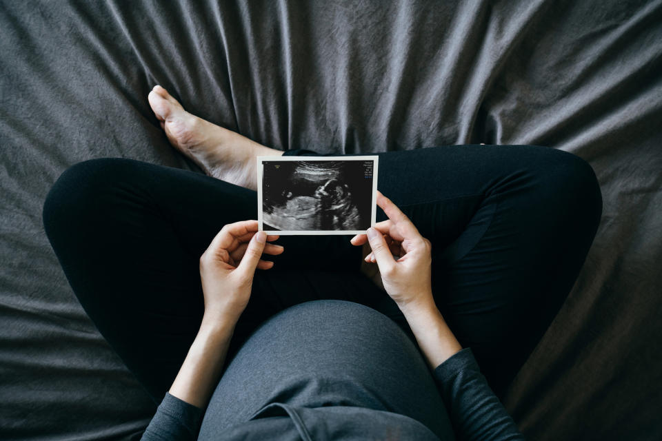 Woman holding an ultrasound picture over a pregnant belly, sitting on a bed