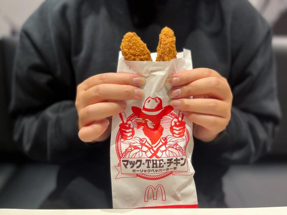 Someone holding a white paper bag with Japanese writing and a picture of a cowboy in red holding two fried chicken tenders.