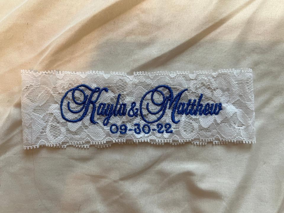 Garter with blue embroidery spelling out "Kayla & Matthew 09-30-22" laid out on a white sheet
