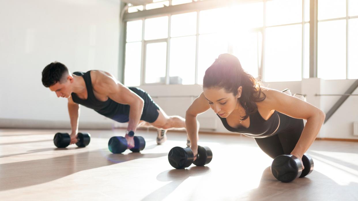  Man and woman side by side performing a push-up using two dumbbells each in a light warehouse gym. 