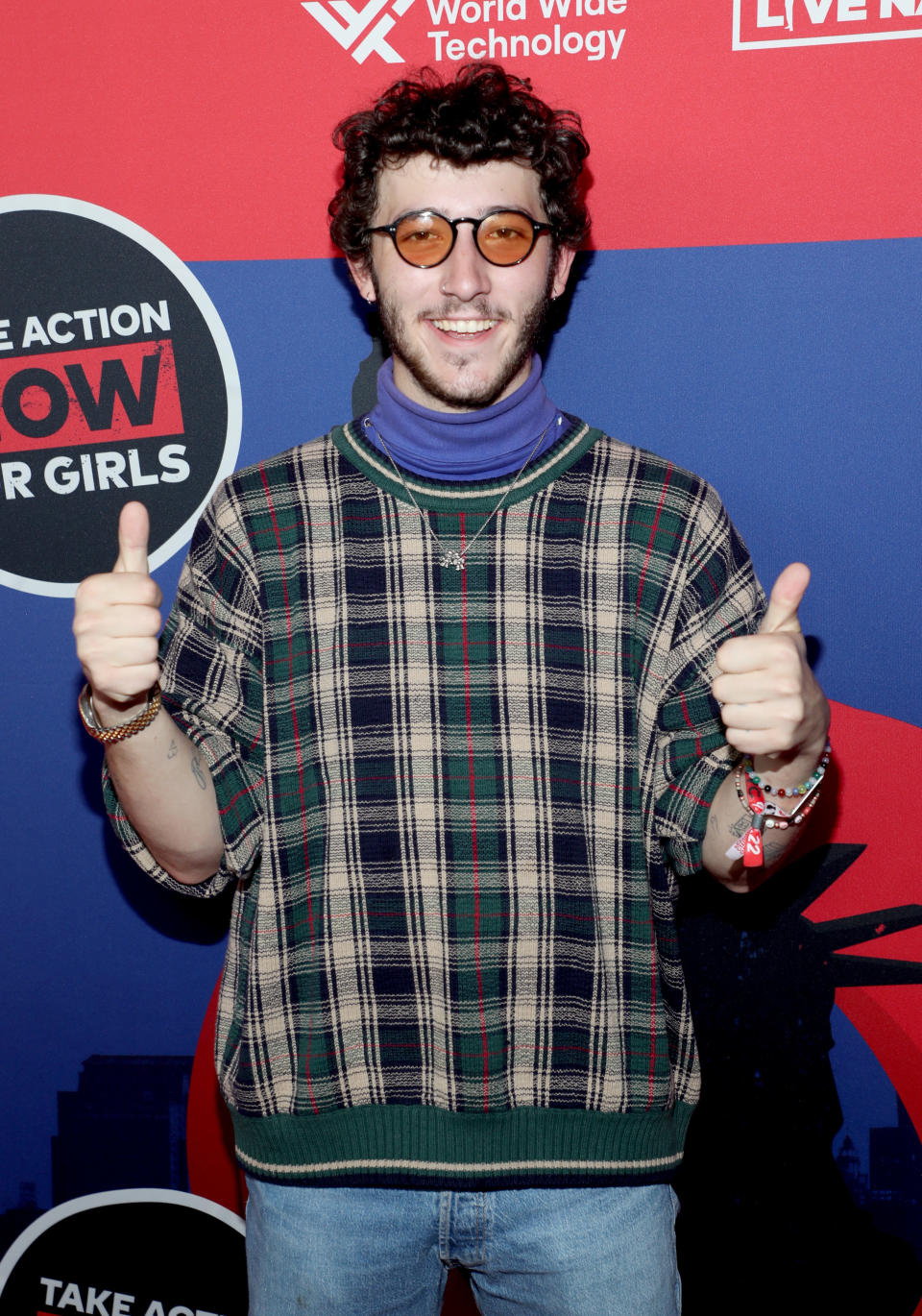 Frankie wearing small circular glasses puts up two thumbs at an event