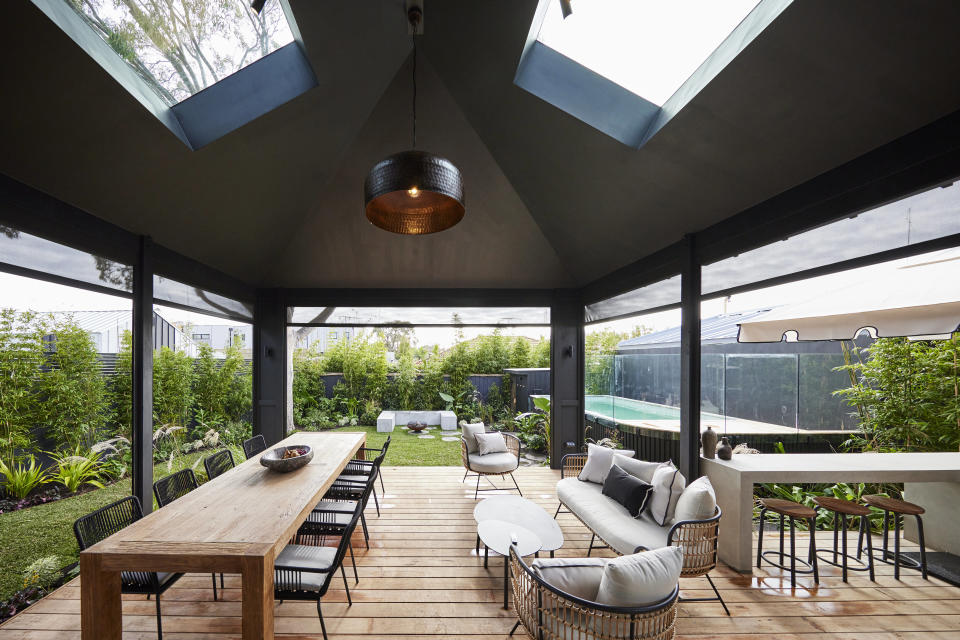 A shot of the deck area with a long table on the left, seats on the right and a black structured ceiling with skylights. 