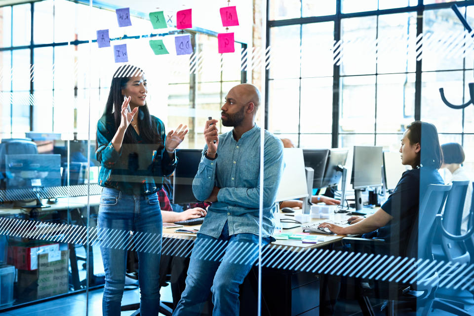 Three colleagues in an office, one standing talking, another listening attentively, third working at a desk