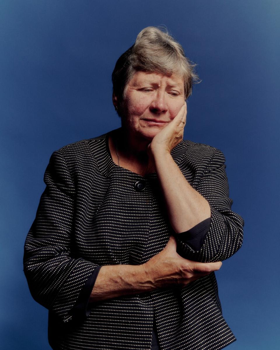 A woman in a gray jacket stands against a blue background with her hand on her face.