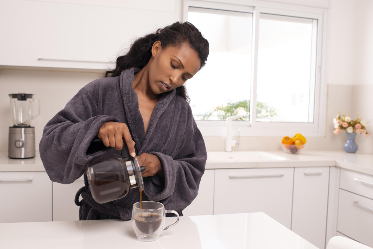 A woman standing in her kitchen wearing a robe pours a cup of coffee.