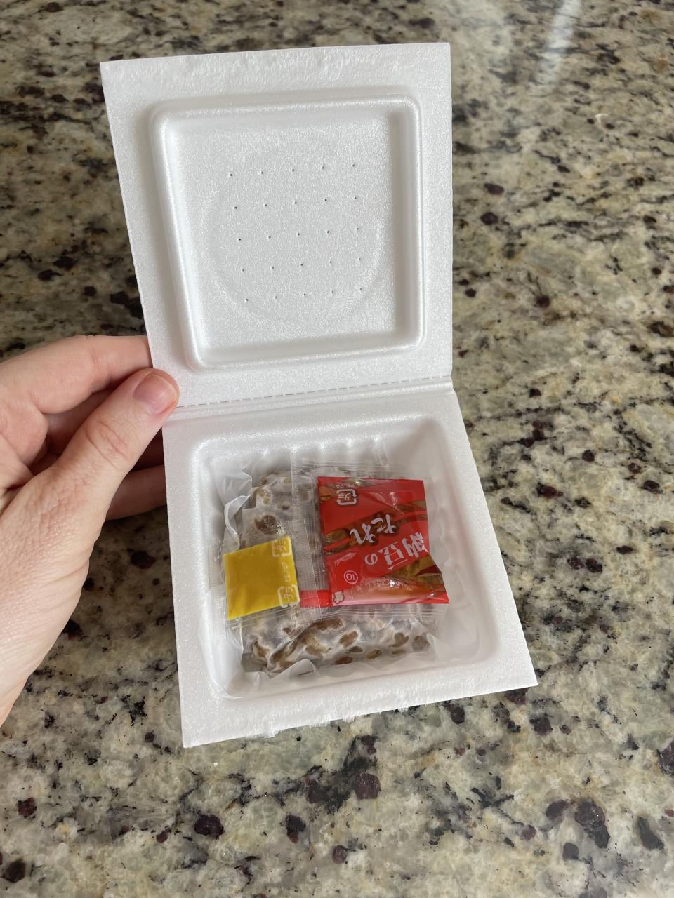 A hand holding an open styrofoam container of natto, with a small packet of sauce and mustard inside