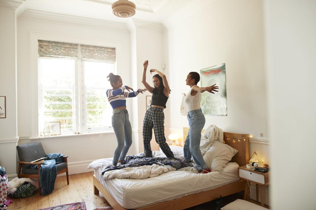 three cheerful young women dancing on bed in modern bedroom with their arms in the air