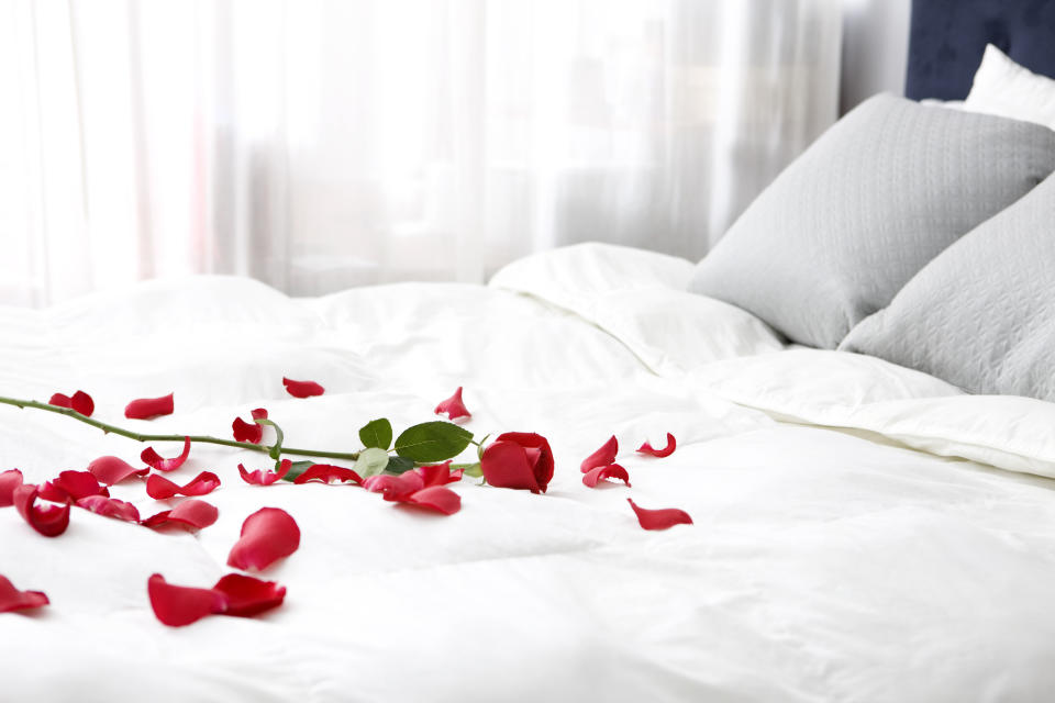 Rose petals scattered on a neatly made bed with grey pillows, suggesting a romantic setting