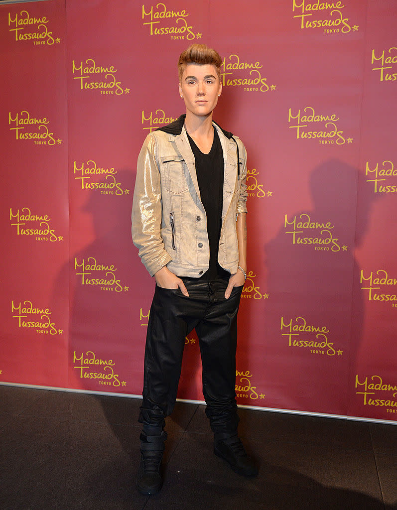 Wax figure of Justin Bieber in a black shirt, black pants, and a light jacket, standing in front of a branded backdrop