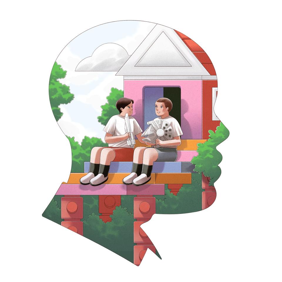 Two children sit in front of a house sharing their lego creations.