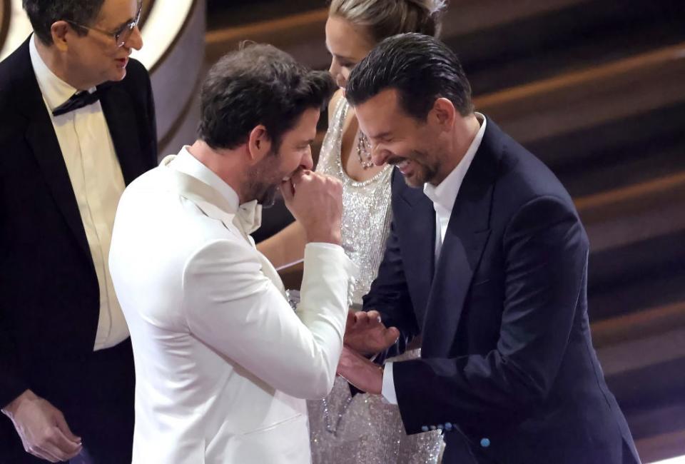 Two men in formal attire sharing a joyful moment onstage, one wearing a white suit and the other in black