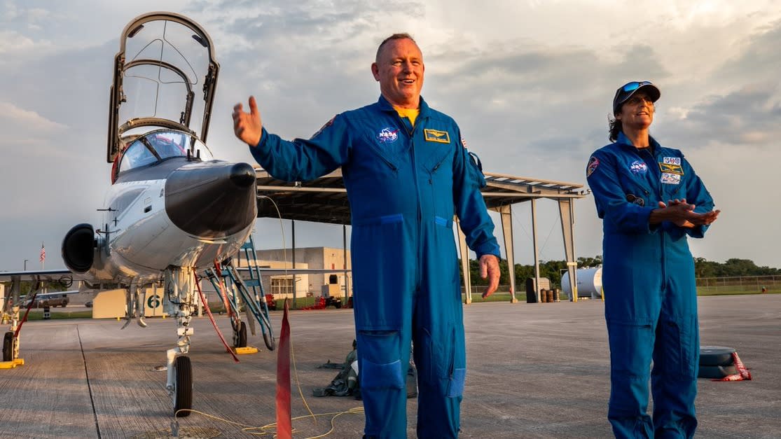  Two astronauts in flight suits standing in front of a t-38 jet with cockpit open. they are on a tarmac with a structure in behind and buildings far in the back. 