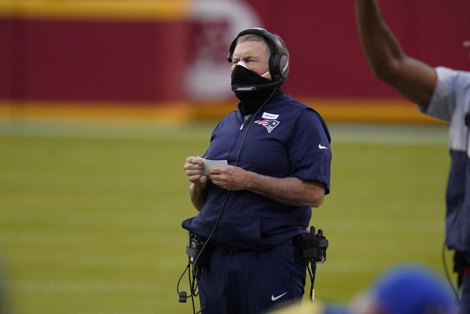 Bill Belichick wears a white face mask covered by a black neck gaiter on the sideline.