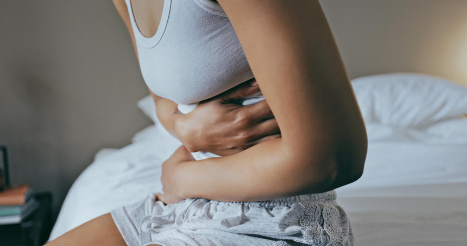 Person sitting on a bed, holding their stomach in pain. They are wearing a tank top and lace-trim shorts