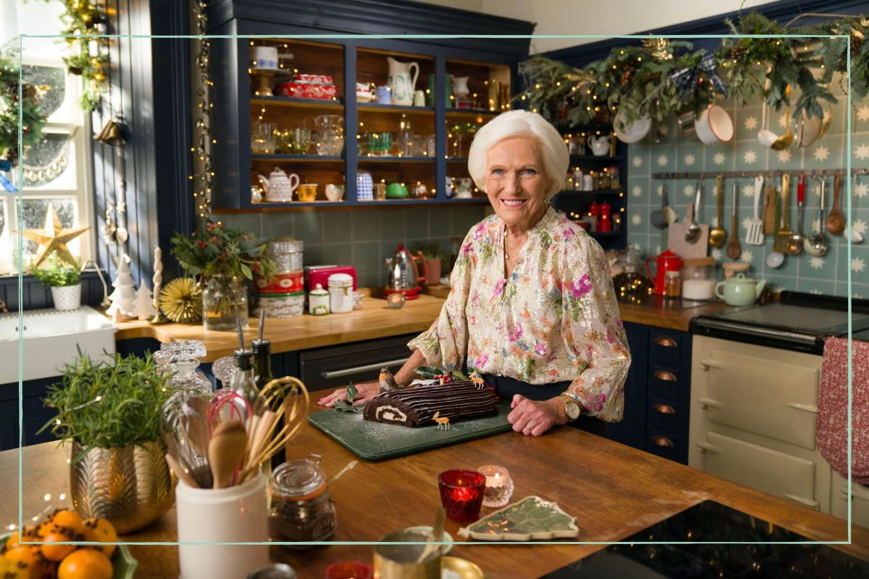  Mary Berry stood next to a Yule Log in a kitchen covered in fairy lights. 