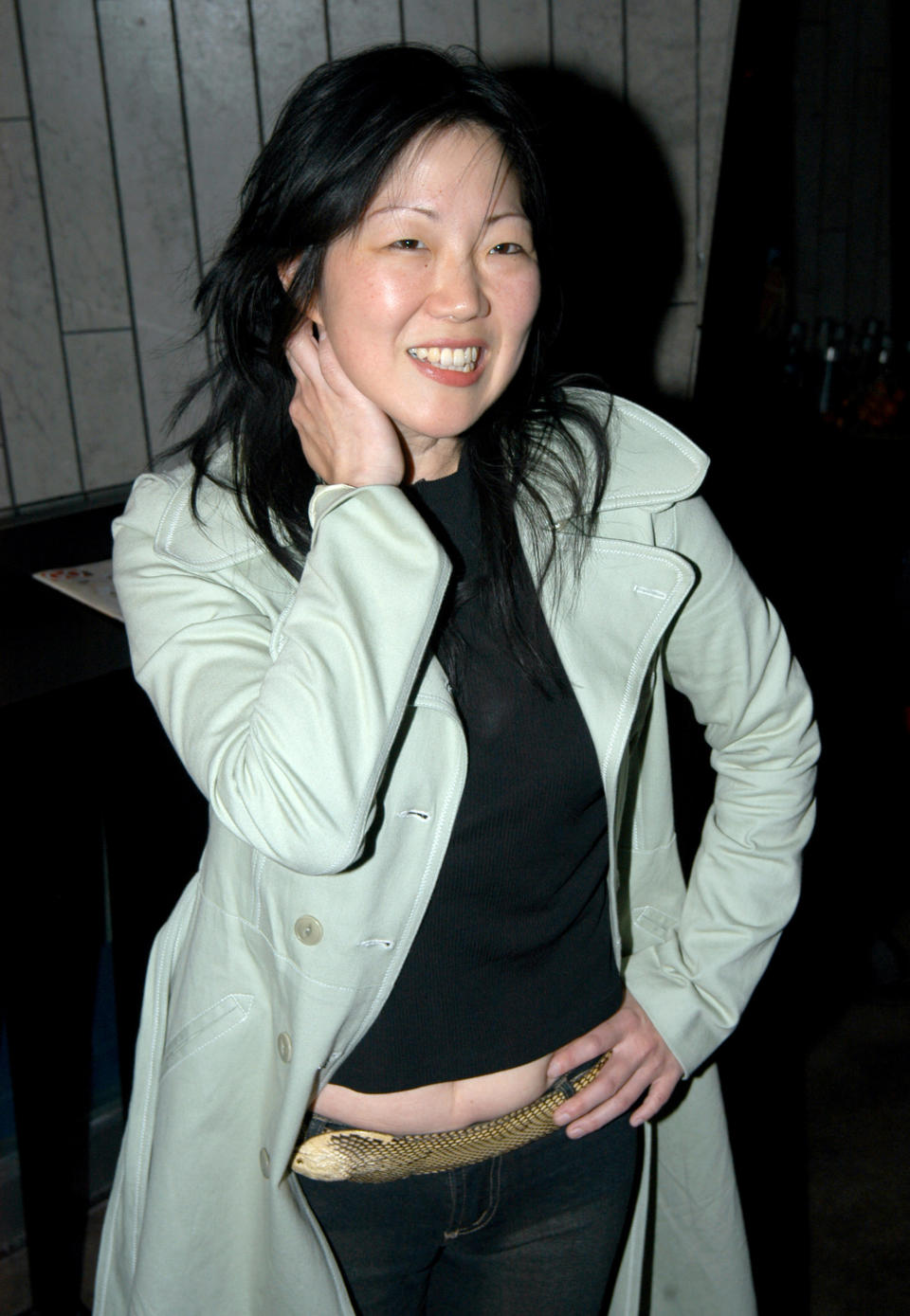 Margaret Cho smiles while posing with one hand on her neck. She is wearing a light trench coat over a black top and dark pants