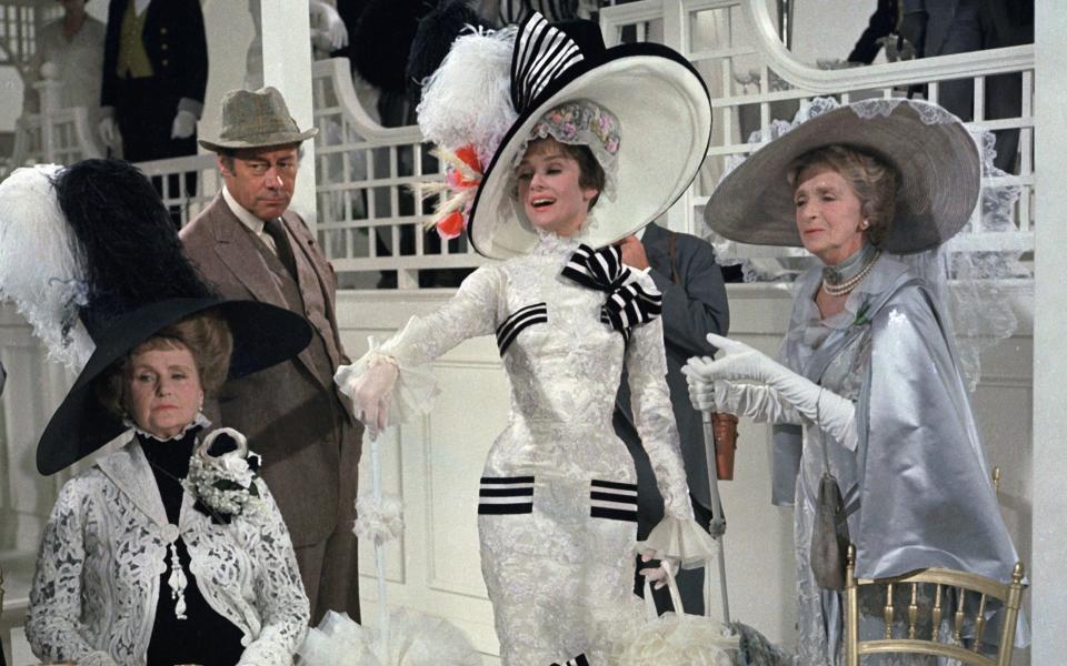 Audrey Hepburn as Eliza Doolittle (center in white dress) and Rex Harrison, (standing atl eft) as Professor Henry Higgins, in "My Fair Lady" - Getty