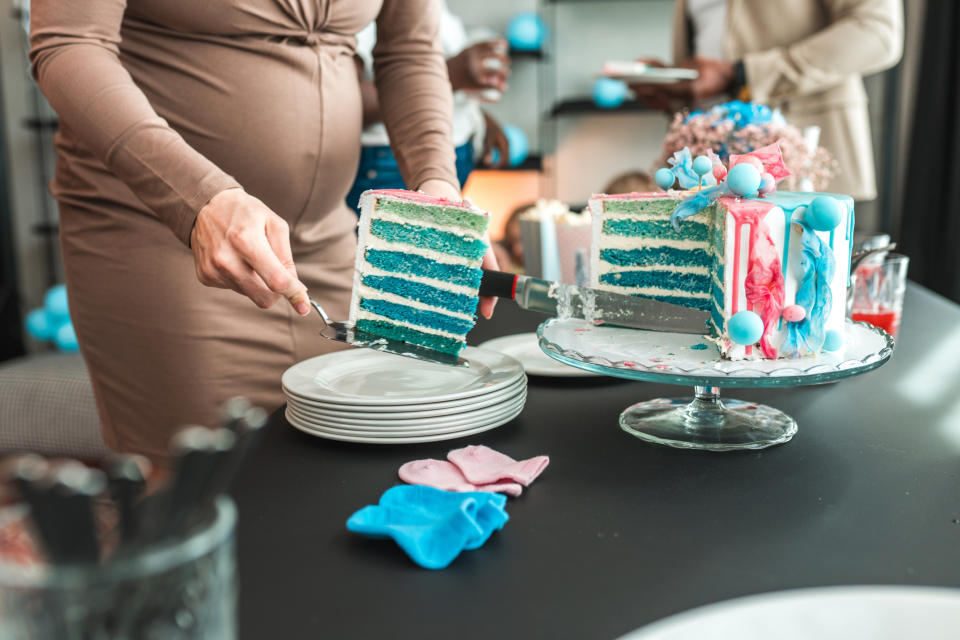 A woman in a beige dress slices a blue and pink layered cake at a party. Other people are in the background. Plates and napkins are on the table