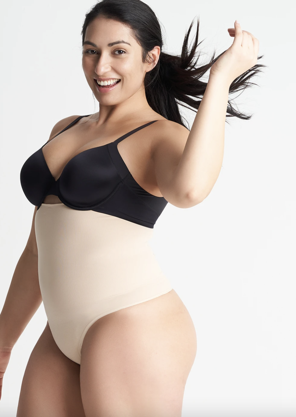 smiling dark hair plus size model wearing a black bra and beige tummy control shapewear